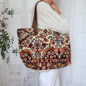 Könnte beinhalten: Eine große Tragetasche mit einem detaillierten floralen und geometrischen Muster im Wandteppich-Stil in den Farben Rot, Schwarz, Creme und Blau. Die Tasche hat zwei braune Leder-Schulterriemen, die mit Metallnieten am oberen Rand befestigt sind.