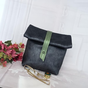 May include: A dark gray clutch with a green strap secured by a silver snap. The clutch is folded over and has a textured appearance. A gold-colored tie clip and a small, round container are in front of the clutch.
