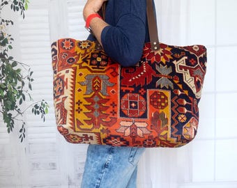 Könnte beinhalten: Eine große Tragetasche mit einem farbenfrohen, gemusterten Design. Die Tasche hat einen Patchwork aus roten, orangefarbenen, gelben und marineblauen geometrischen Formen. Sie hat braune Ledergriffe. Eine Person trägt die Tasche, bekleidet mit einem blauen Oberteil und Jeans.