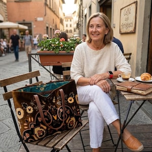 May include: A large brown velvet tote bag featuring a dark floral pattern with cream, orange, and black accents. The bag has two brown leather handles and a teal interior lining with a visible floral print, resting on a wooden chair.