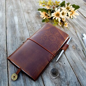 May include: A brown leather journal with a decorative gear pattern and a silver pen. The journal has an elastic band closure and a brass button.