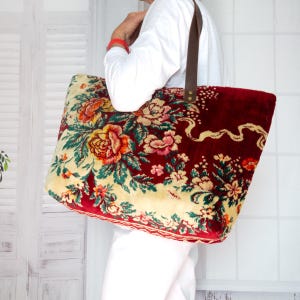 May include: A large tote bag crafted from plush, deep red velvet fabric featuring a prominent floral pattern in shades of orange, yellow, and green. The bag includes dark brown leather shoulder straps secured with metallic rivets at the top edge.