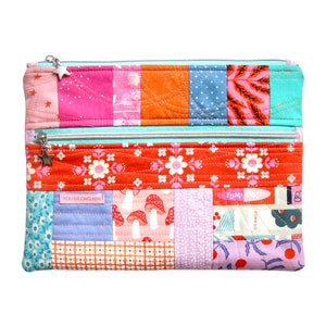 Puede incluir: Una bolsa de cremallera de tela patchwork con dos compartimentos. La bolsa est&aacute; hecha de varias telas coloridas con patrones florales, de hongos y geom&eacute;tricos. La bolsa tiene dos tiradores de cremallera plateados con abalorios en forma de estrella. El texto "YOU BELONG HERE!" y "YUM!" es visible en la bolsa.
