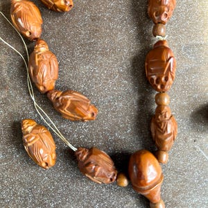 May include: Close-up of a wooden beaded necklace and loose beads. The beads are carved in a detailed insect design, with a warm brown color. The necklace has a tassel at the end. The beads are strung on a thin, light-colored string.