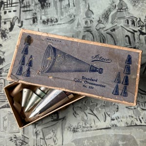 May include: An open cardboard box containing several silver metal cake decorating tips. The box is labeled "Ateco Standard Cake Ornamentor No. 333" with a diagram of various tip shapes. The box is blue and tan with the text "Made in U.S.A."