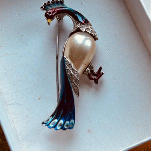 May include: A vintage peacock brooch with a faux pearl body, blue and red enamel, and rhinestones. The peacock's tail feathers are blue with white eyes.