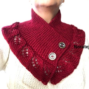 May include: A red knitted cowl with a scalloped edge and two buttons. The cowl is worn by a person with a white sweater underneath.