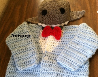 Puede incluir: Un cárdigan azul de ganchillo para bebé con una corbata de lazo blanca de ganchillo y un gorro de tiburón gris de ganchillo con ojos negros.