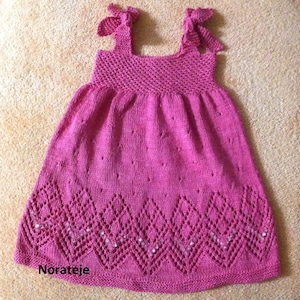 May include: A pink knitted baby dress with a lace pattern and a diamond-shaped design at the bottom. The dress has straps with bows and is decorated with small silver beads.