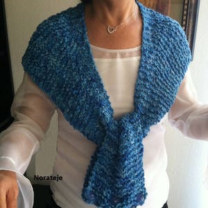 May include: A blue knitted scarf with a textured pattern and a knotted design. The scarf is worn by a person with a white shirt and a silver necklace with a heart pendant.