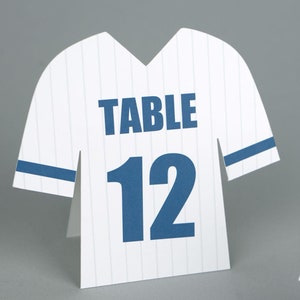 May include: White baseball jersey-shaped table number cards with blue stripes and text. The cards feature the word "TABLE" above the number "12". The design is reminiscent of a baseball uniform.