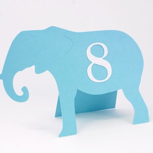 May include: A light blue paper elephant cutout with a white number 8 in the center. The elephant has a curled trunk and is standing on four legs. The cutout is likely a decorative item or a table centerpiece.