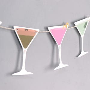 May include: A string of paper martini glasses in various colors, including pink, green, and brown, strung on a natural twine. Each glass has a white stem and is decorated with a lemon twist or olive. The background is a solid gray.