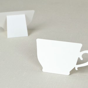 Puede incluir: Dos recortes de tazas de té de papel blanco, uno de pie y el otro tumbado de lado. Las tazas de té tienen una forma clásica con un asa curvada. El fondo es una tela beige claro.