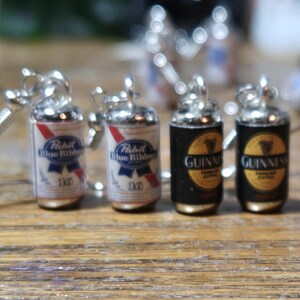 Puede incluir: Cuatro pendientes de cerveza en miniatura, dos son azules y rojos con el texto "Pabst Blue Ribbon 12oz" y dos son negros con el texto "Guinness Foreign Extra Stout"