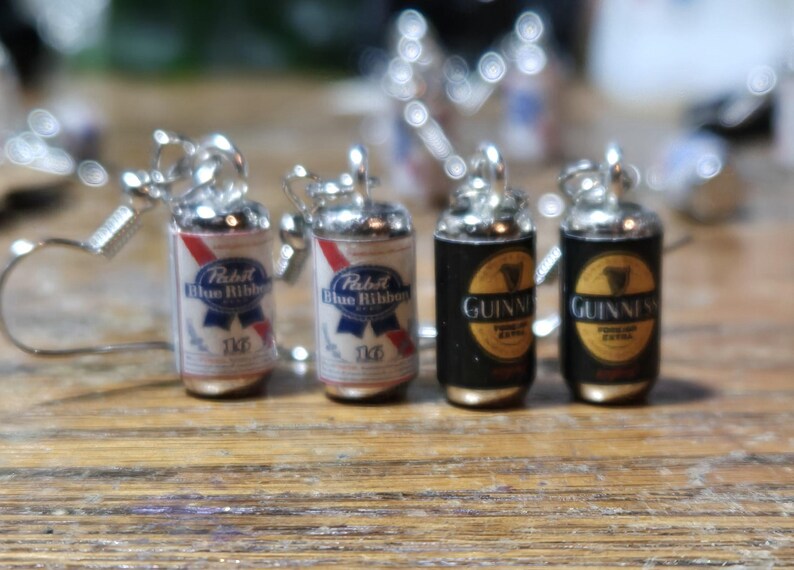Puede incluir: Cuatro pendientes de cerveza en miniatura, dos son azules y rojos con el texto "Pabst Blue Ribbon 12oz" y dos son negros con el texto "Guinness Foreign Extra Stout"