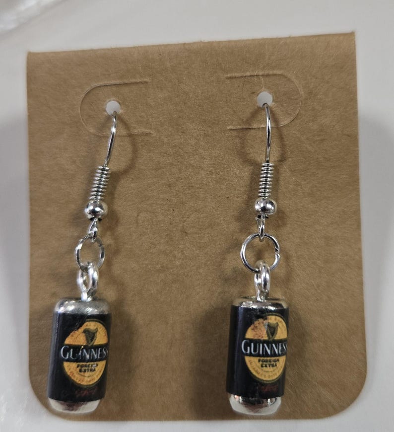 Puede incluir: Un par de pendientes de aro de plata con un encanto de lata de Guinness en miniatura. La lata es negra con una etiqueta dorada que dice "Guinness Foreign Extra Stout".