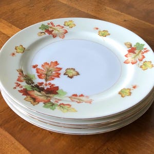 May include: A stack of four white ceramic plates with a delicate autumn leaf design in shades of red, orange, and green. The plates have a gold rim and a light cream-colored border. The plates are stacked on a wooden surface.