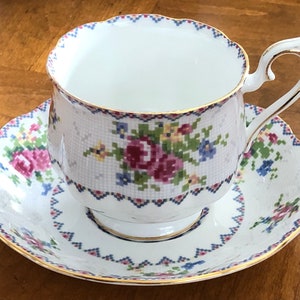 May include: A white teacup and saucer with a floral pattern. The pattern features pink, blue, and green flowers on a white background. The cup and saucer have a gold trim.