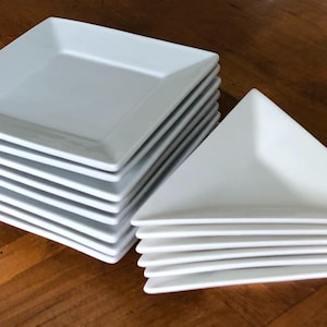 May include: A collection of white square and triangular plates stacked on a wooden surface. The plates have a clean, minimalist design, suitable for serving appetizers or desserts. The square plates are stacked in two piles, while the triangular plates are stacked in a separate pile.