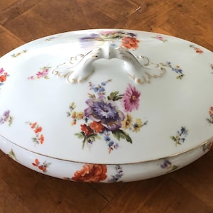 May include: White oval porcelain serving dish with a lid. The dish is decorated with a floral pattern in shades of pink, purple, orange, and yellow. The lid has a gold-painted handle.