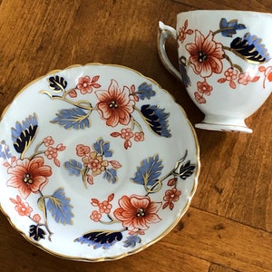 May include: A white teacup and saucer with a floral pattern in shades of blue, red, and gold. The floral design features stylized leaves and flowers.