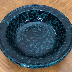 May include: A round, black bowl with a speckled teal pattern. The bowl has a wide rim and a glossy finish. The interior of the bowl also features the teal speckled design. The bowl is sitting on a wooden surface.