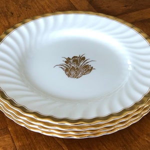 May include: A stack of four white ceramic dinner plates with a gold rim and a gold floral design in the center.