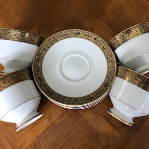 May include: A set of white porcelain teacups and saucers with gold floral detailing. The teacups have a unique angled design and sit on small pedestal bases. The saucers and the rims of the teacups are adorned with intricate gold patterns.