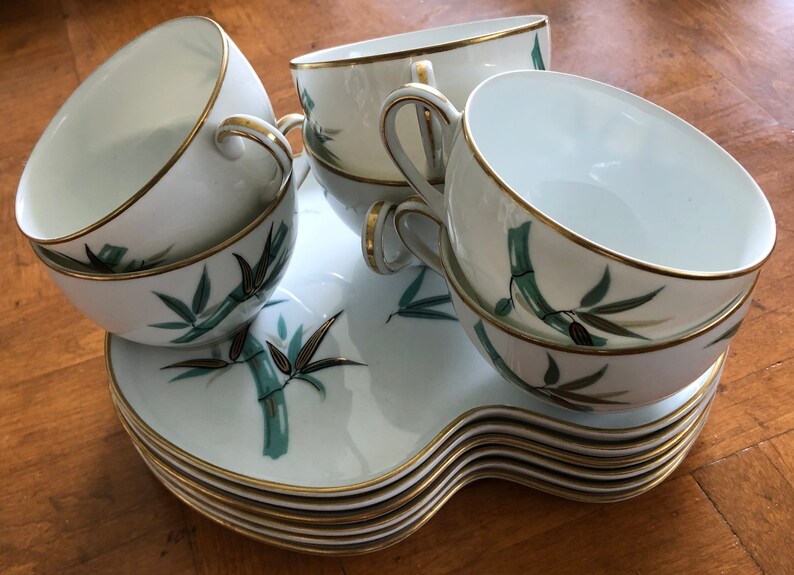 May include: A set of white teacups and saucers with a green bamboo design and gold trim. The cups and saucers are stacked on a white tray with a gold trim.