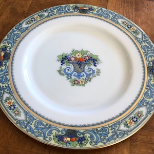 May include: A white ceramic dinner plate with a blue and gold floral border. The center of the plate features a basket of fruit with grapes, apples, and pears.