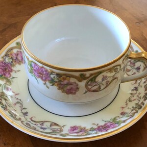 Puede incluir: Una taza de t&eacute; y un platillo blancos con un patr&oacute;n floral en tonos de rosa, verde y dorado. La taza y el platillo tienen un borde dorado y una banda negra alrededor del borde.