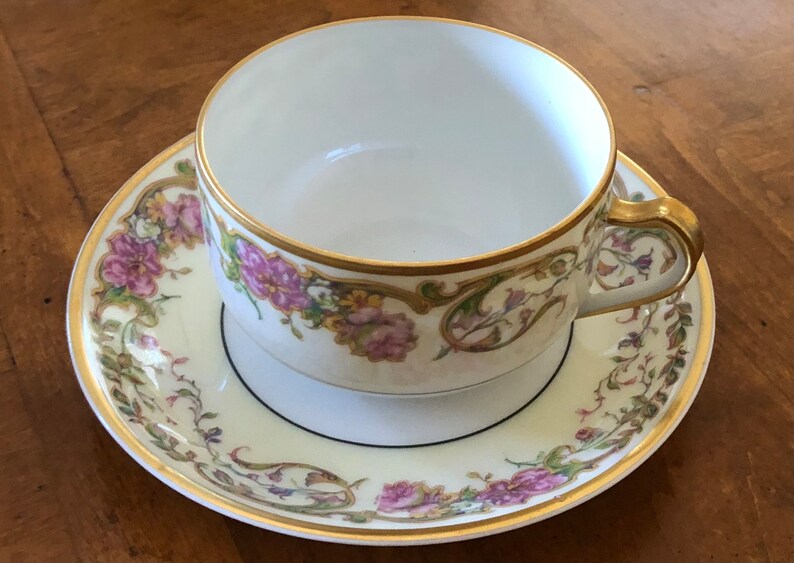 Puede incluir: Una taza de t&eacute; y un platillo blancos con un patr&oacute;n floral en tonos de rosa, verde y dorado. La taza y el platillo tienen un borde dorado y una banda negra alrededor del borde.