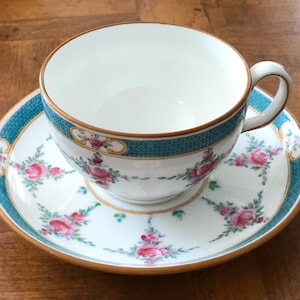 May include: A white teacup and saucer set with a floral design. The cup and saucer have a teal border with a gold trim. Pink roses and green leaves are featured on the saucer and cup.