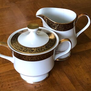 May include: White porcelain sugar bowl with lid and creamer. Both pieces have gold trim and a decorative brown floral pattern. The sugar bowl has a gold handle and a gold knob on the lid. The creamer has a curved spout and a gold handle.