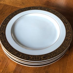 May include: A stack of four white ceramic plates with a gold rim and a decorative brown floral border. The plates are stacked on top of each other, showcasing the design.