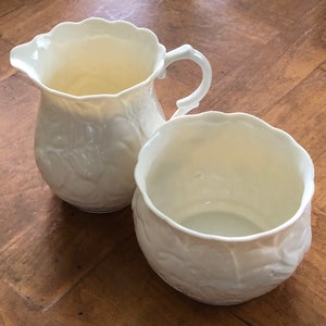 May include: Two white ceramic creamer and sugar bowls with an embossed floral design.