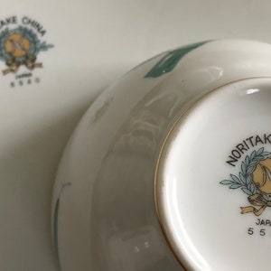 May include: Close-up of the bottom of a white ceramic bowl with a green floral design. The bowl has a gold rim and a Noritake China logo with the text "Japan 5540".