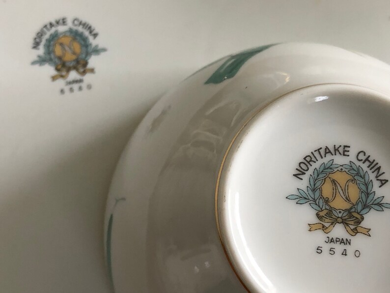 May include: Close-up of the bottom of a white ceramic bowl with a green floral design. The bowl has a gold rim and a Noritake China logo with the text "Japan 5540".