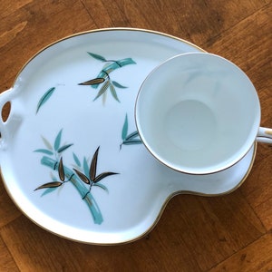 May include: A white ceramic teacup and saucer set with a gold rim and a bamboo design. The saucer has a unique shape with a handle on the side.