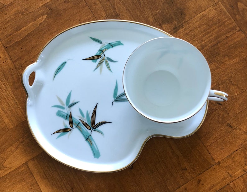 May include: A white ceramic teacup and saucer set with a gold rim and a bamboo design. The saucer has a unique shape with a handle on the side.