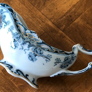 May include: A white porcelain gravy boat with a blue and gold floral design. The boat has a handle and a spout.