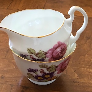 May include: A white porcelain creamer and sugar bowl set. The set features a floral design with pink roses, purple flowers, and green leaves. The rims are trimmed with gold. The creamer has a handle and a spout.