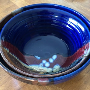 May include: Nested ceramic bowls with a glossy finish. The interior bowl is a deep blue, with a band of red and white accents. The outer bowl is a darker blue with a brown rim. The bowls are on a wooden surface.