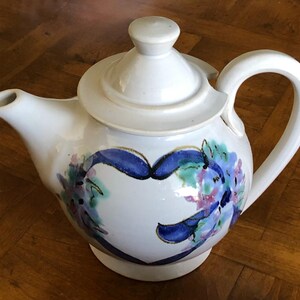 May include: White teapot with a blue and green floral design and a heart shape in the center. The teapot has a lid and a handle.