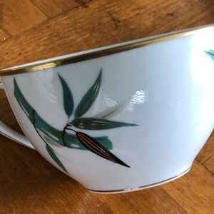 May include: A white porcelain teacup with a gold rim and a green and gold bamboo design.