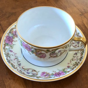 Puede incluir: Una taza de t&eacute; y un platillo blancos con un patr&oacute;n floral en tonos de rosa, verde y dorado. La taza y el platillo tienen un borde dorado y una l&iacute;nea negra alrededor del borde.