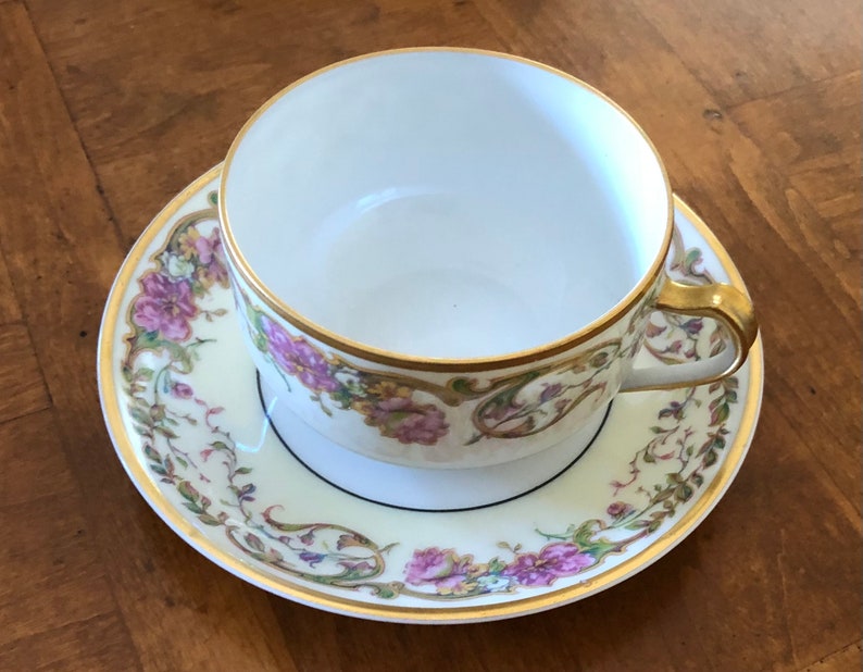 Puede incluir: Una taza de t&eacute; y un platillo blancos con un patr&oacute;n floral en tonos de rosa, verde y dorado. La taza y el platillo tienen un borde dorado y una l&iacute;nea negra alrededor del borde.