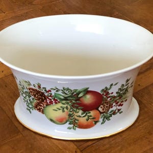 May include: White ceramic planter with a wide, shallow bowl shape. The planter features a colorful fruit and foliage design around the base, including apples, pears, and pinecones. A gold trim accents the rim and base.