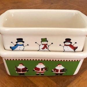 May include: Two stacked rectangular ceramic baking dishes. The top dish features three snowmen with hats and scarves. The bottom dish has a green border with three Santas. The dishes are off-white.